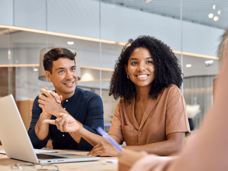 Two people smiling and sitting in a modern office with a laptop open in front of them.