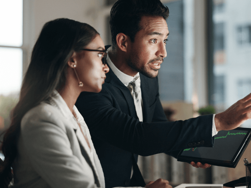 Two professionals discussing while looking at a tablet with a graph in an office setting.