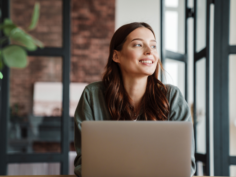 A woman smiling and looking away from her laptop in a bright office.