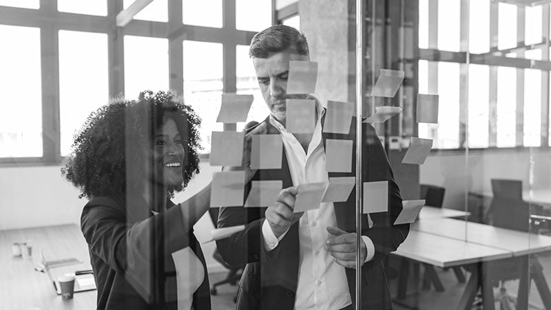 Two consultants collaboratively mapping a customer journey strategy using colorful sticky notes on a glass wall in a modern office.