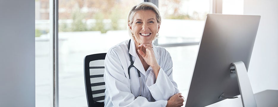A smiling woman in a white coat sits at a computer, engaged in her work.