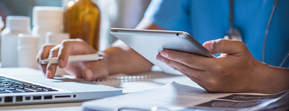 A doctor is focused on using a laptop while holding a tablet in their other hand.