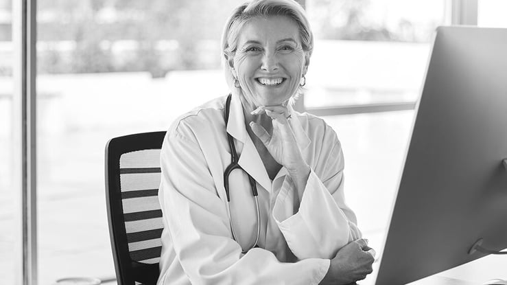 A smiling woman in a white coat sits at a computer, engaged in her work.