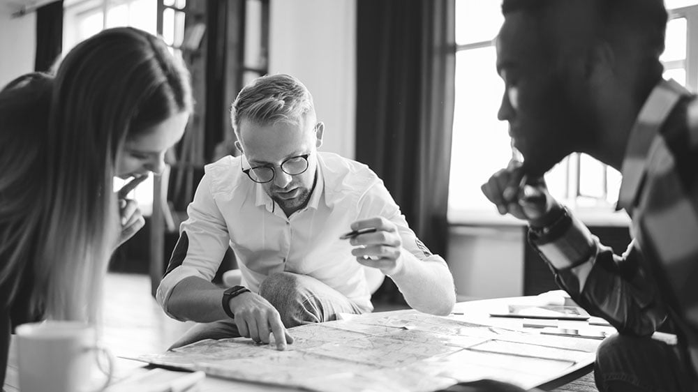 A diverse consulting team huddles over a strategic roadmap, collaboratively identifying key touchpoints to improve the client's customer experience.