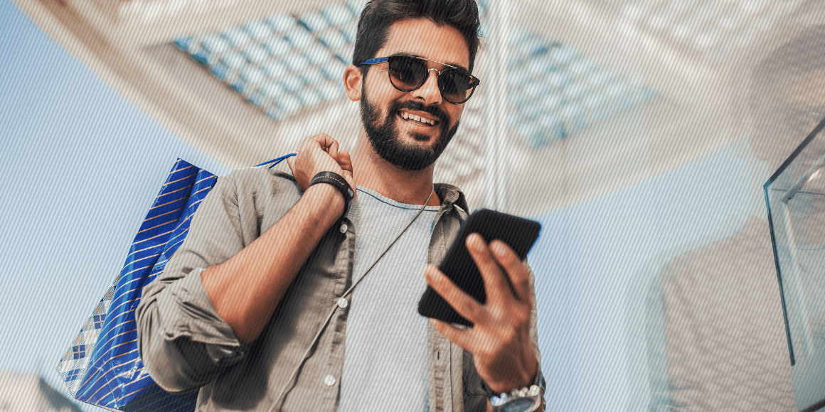 Featured image: Young man walking out of a store holding shopping bags and using their mobile phone - Read full post: How Has Hybrid Shopping Affected VoC